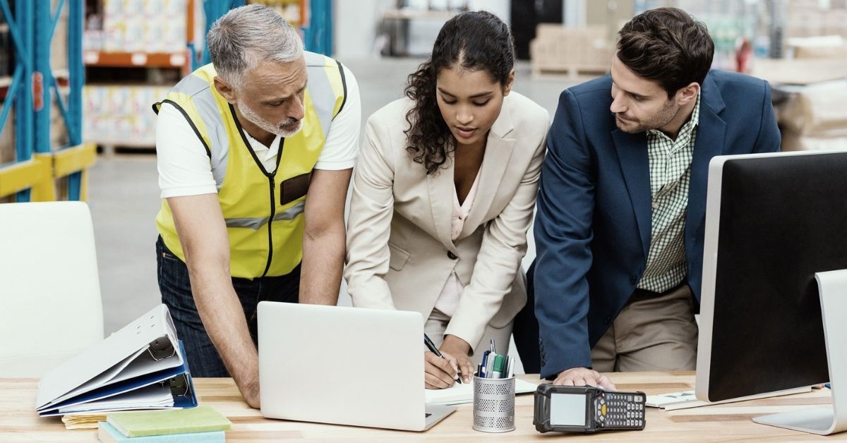 Three people collaborating over a laptop and documents in a warehouse setting.